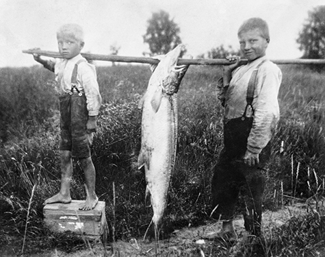 Two young boys wearing suspenders hold up their catch of fish.
