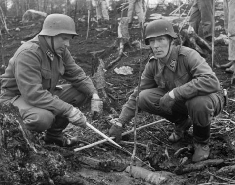 Two men dressed in army uniforms are handling an ordnance on the ground in a forested terrain.