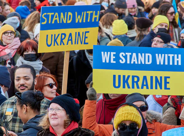 A group of people with signs. Signs painted with yellow and blue include texts: STAND WITH UKRAINE